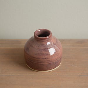 May include: A small, round, handmade ceramic vase with a narrow neck. The vase is a muted reddish-purple color with darker swirling patterns. The base is a lighter, unglazed color. The vase is sitting on a wooden surface.