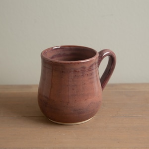 Può includere: Una tazza in ceramica fatta a mano con un corpo arrotondato e un manico. La tazza è di un colore rosso-viola intenso con sottili variazioni di tonalità. La base è di un colore bianco sporco più chiaro. La tazza è su una superficie di legno.