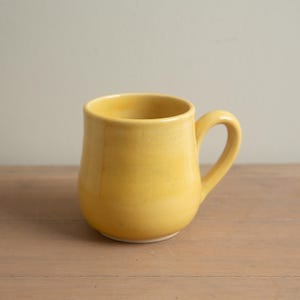 Può includere: Una tazza in ceramica gialla con un corpo arrotondato e un manico curvo. La tazza ha una finitura liscia e lucida. La tazza è vuota e pronta per essere riempita con una bevanda. Si trova su una superficie di legno.
