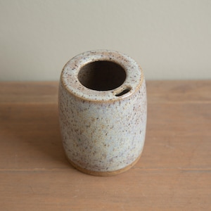 Può includere: Un portaspazzolino in ceramica macchiata. Il supporto cilindrico ha un'ampia apertura nella parte superiore e un piccolo foro sul lato. Il colore è un mix di beige, marrone e azzurro, con una superficie strutturata. La base è marrone pieno.