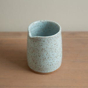 Può includere: Brocca in ceramica azzurra maculata con beccuccio. La brocca ha un corpo arrotondato e una base naturale non smaltata. L'interno è dello stesso colore azzurro dell'esterno. Posizionata su una superficie di legno.