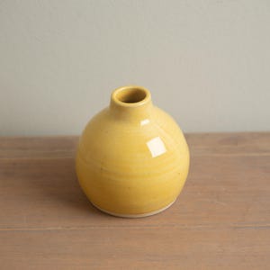 Może przedstawiać: Mały, okrągły, żółty ceramiczny wazon z wąską szyjką. Wazon ma gładkie, błyszczące wykończenie i stoi na drewnianej powierzchni. Wazon jest przedmiotem dekoracyjnym, odpowiednim do trzymania kwiatów lub jako samodzielny element.