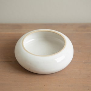 May include: A small, round, white ceramic bowl with a wide opening. The bowl has a smooth, glossy exterior and a slightly textured interior rim. The bowl is sitting on a wooden surface.