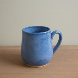 May include: A handmade blue ceramic mug with a rounded body and a curved handle. The mug has a speckled texture and a glossy finish. The base of the mug is a light beige color. The mug is designed for hot beverages.