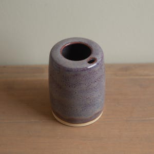 May include: A cylindrical ceramic vase in shades of purple with a speckled texture. The vase has a wide opening at the top and a smaller hole on the side. The base is a lighter colour, and the rim has a darker accent.