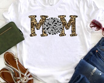 May include: White t-shirt with the word "MOM" in black letters with a leopard print background. The "O" is replaced with a white and gray cheerleading pom-pom with a red heart in the center.