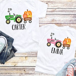 May include: Two white t-shirts with a green and pink tractor pulling a wagon full of pumpkins. The shirts are personalized with the names "Carter" and "Emma".