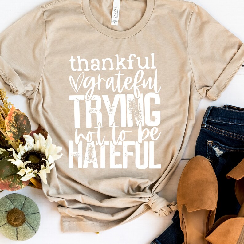 Puede incluir: Una camiseta beige con el texto "thankful grateful trying not to be hateful" impreso en blanco.