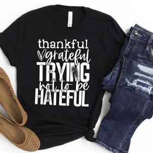 Puede incluir: Camiseta negra con texto blanco que dice "thankful grateful trying not to be hateful". La camiseta est&aacute; sobre una superficie blanca con un par de pantalones vaqueros azules y sandalias marrones.