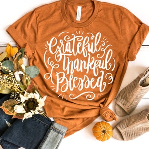 May include: A brown t-shirt with the text "Grateful & Thankful Blessed" in white lettering. The shirt is laid out on a white surface with a small orange pumpkin, a pair of brown sandals, and a bouquet of fall flowers.
