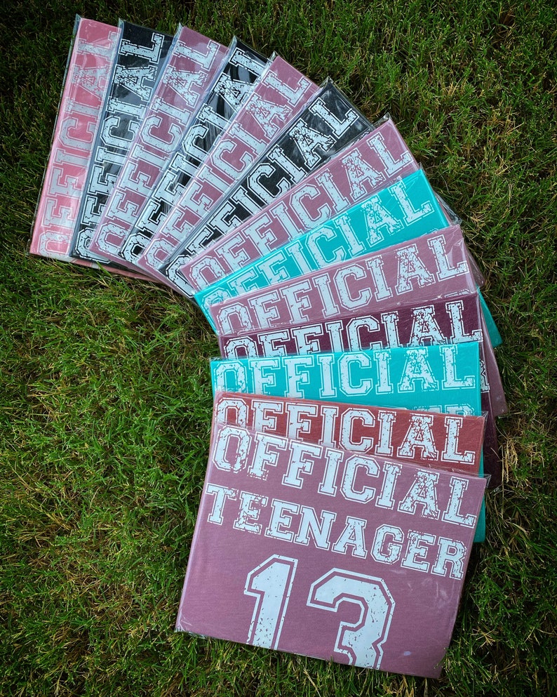 May include: A stack of maroon, teal and black t-shirts with the text "OFFICIAL TEENAGER 13" printed on the front.