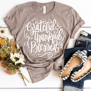 May include: A gray t-shirt with the text "Grateful Thankful Blessed" in white lettering. The shirt is folded and tied in a knot. The shirt is on a white surface with a bouquet of flowers and a pair of jeans.
