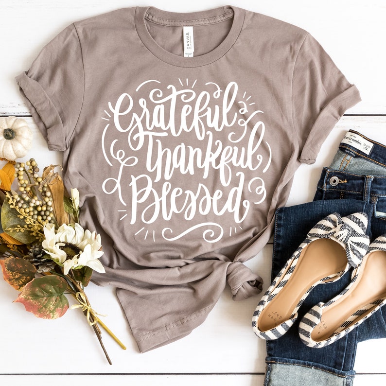 May include: A gray t-shirt with the text "Grateful Thankful Blessed" in white lettering. The shirt is folded and tied in a knot. The shirt is on a white surface with a bouquet of flowers and a pair of jeans.