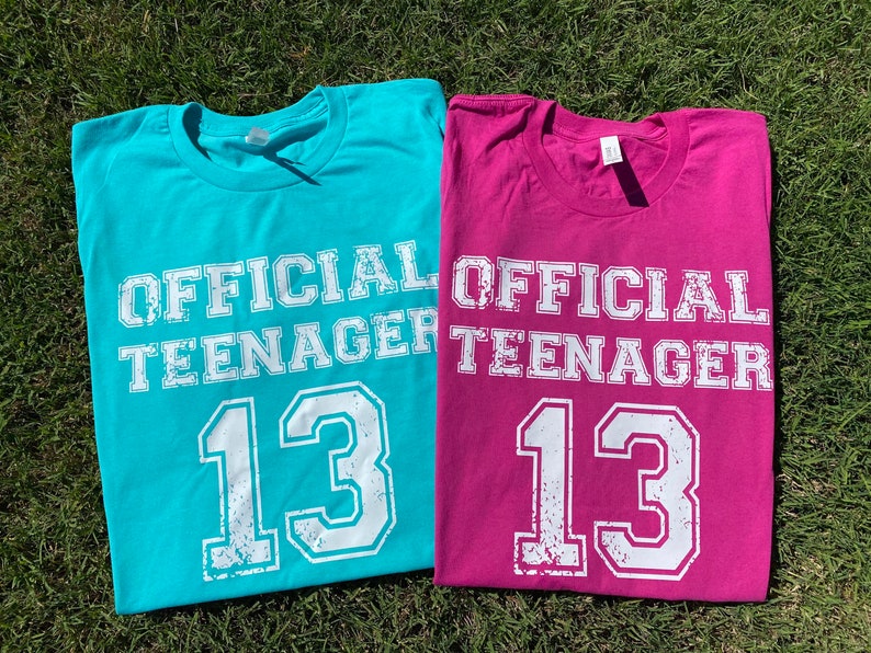 May include: Two t-shirts, one turquoise and one pink, with the text "OFFICIAL TEENAGER 13" in white.
