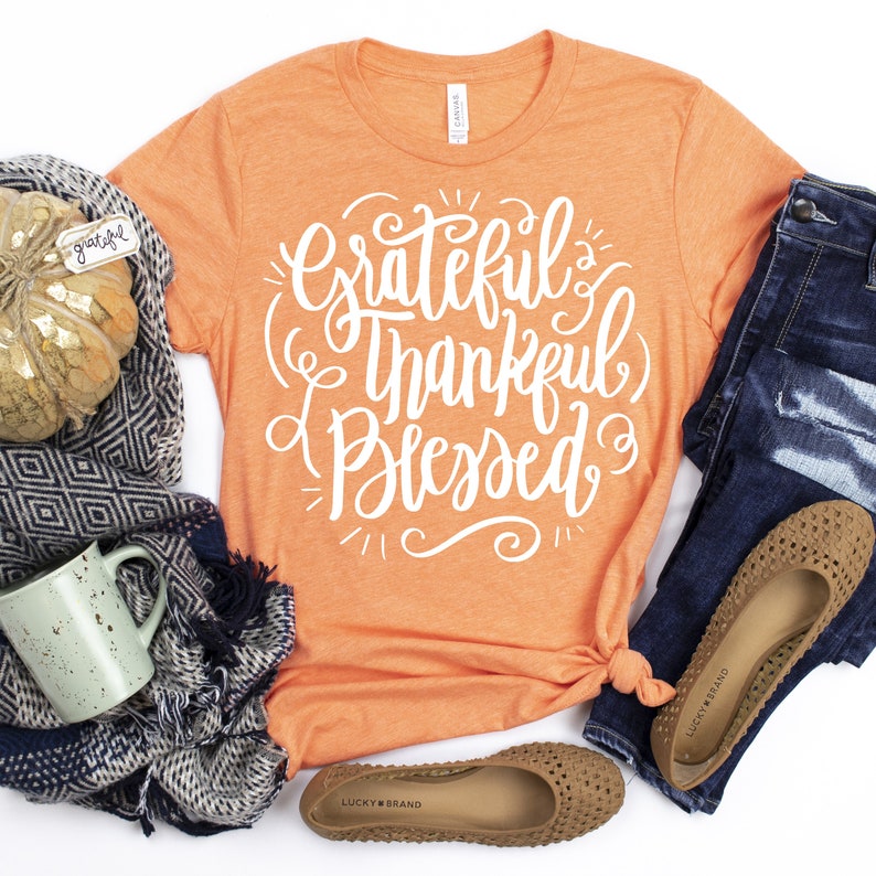 May include: Orange t-shirt with the text "Grateful Thankful Blessed" in white lettering. The shirt is styled with a scarf, a mug, and brown flats.