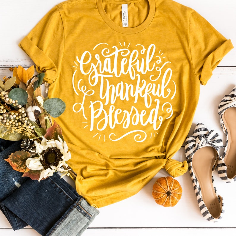 May include: A mustard yellow t-shirt with the text "Grateful Thankful Blessed" in white lettering. The shirt is laid flat on a white surface with a small orange pumpkin, a pair of black and white striped flats, and a bouquet of fall flowers.