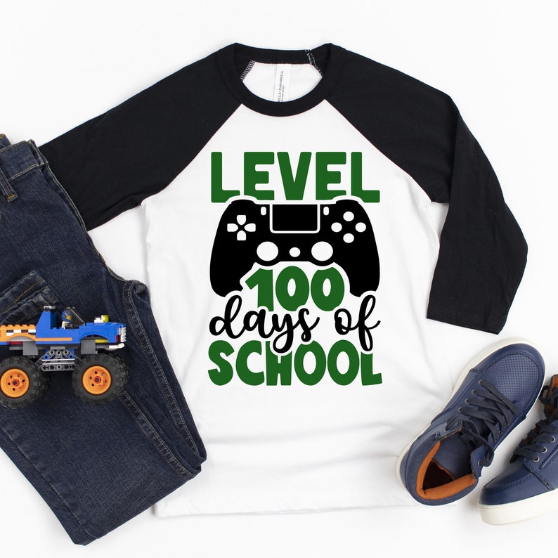 May include: A white and black baseball tee shirt with green text that reads "Level 100 days of school" and a black video game controller graphic.