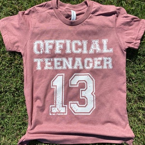 May include: A maroon t-shirt with the text "OFFICIAL TEENAGER 13" in white distressed lettering.