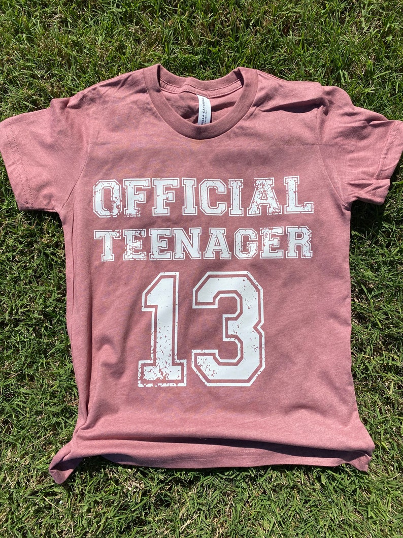 May include: A maroon t-shirt with the text "OFFICIAL TEENAGER 13" in white distressed lettering.