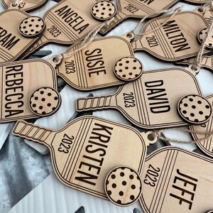 Puede incluir: Adornos navide&ntilde;os personalizados de madera en forma de paleta, cada uno grabado con un nombre y un a&ntilde;o. Un peque&ntilde;o adorno redondo con forma de galleta y lunares marr&oacute;n oscuro acompa&ntilde;a a cada paleta. Los nombres incluyen Kerynne, Angela, Susie, Kristen y otros.