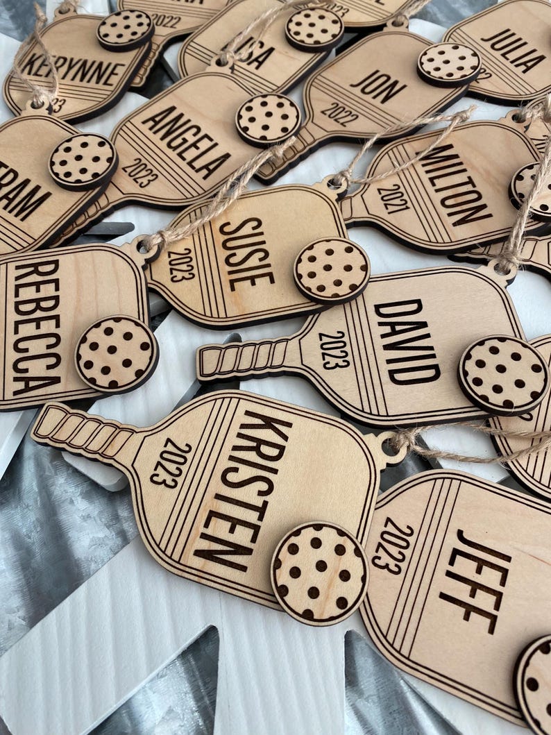 Puede incluir: Adornos navide&ntilde;os personalizados de madera en forma de paleta, cada uno grabado con un nombre y un a&ntilde;o. Un peque&ntilde;o adorno redondo con forma de galleta y lunares marr&oacute;n oscuro acompa&ntilde;a a cada paleta. Los nombres incluyen Kerynne, Angela, Susie, Kristen y otros.