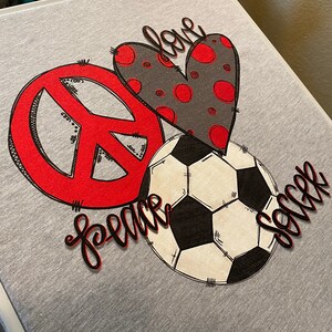 May include: A gray t-shirt with a red peace sign, a black and white soccer ball, and a red and gray heart with the word "love" written on it. The words "peace" and "soccer" are written in black cursive.