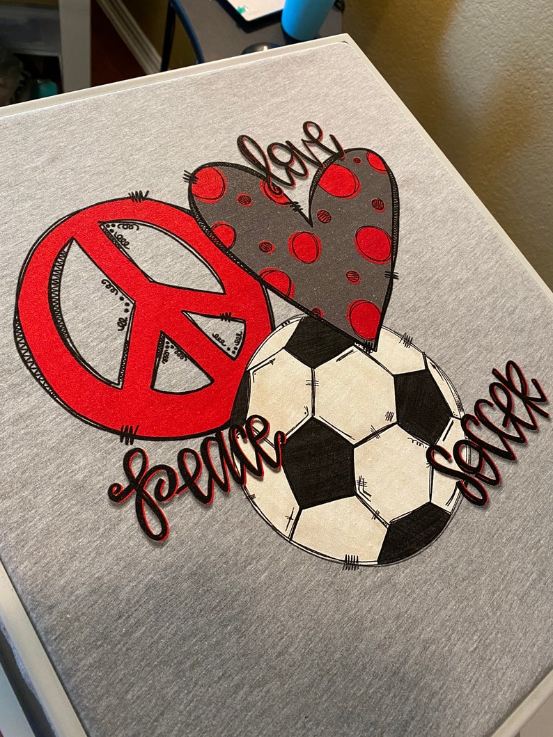 May include: A gray t-shirt with a red peace sign, a black and white soccer ball, and a red and gray heart with the word "love" written on it. The words "peace" and "soccer" are written in black cursive.