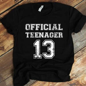 May include: Black t-shirt with white text that reads "OFFICIAL TEENAGER 13".