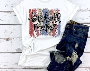 May include: White t-shirt with a distressed graphic design featuring a baseball, red, white, and blue stripes, and leopard print. The text "baseball mama" is written in black cursive lettering.