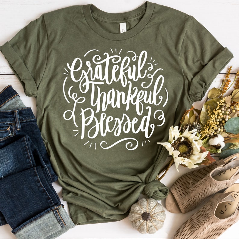 May include: A green t-shirt with the text "Grateful Thankful Blessed" in white lettering. The shirt is folded and displayed with a pair of jeans and sandals.