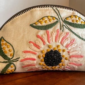 May include: A half-moon shaped pouch made of off-white fabric featuring hand-embroidered floral designs in pink, yellow, green, and dark blue. The pouch includes a dark metal zipper closure and a light tan leather pull tab attached to the side.