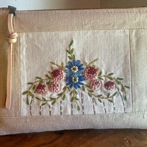 May include: A rectangular beige fabric pouch featuring a central embroidered floral design with blue and pink flowers, green leaves, and thin vertical grey stitched lines. A light tan leather pull tab is attached to the metal zipper closure.