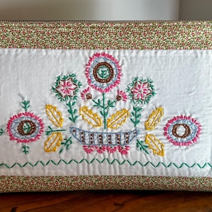 May include: A rectangular zippered pouch featuring a central white panel with colorful floral embroidery in pink, yellow, and green. The pouch is bordered by a green fabric with a small red and white floral print and includes a light leather zipper pull.