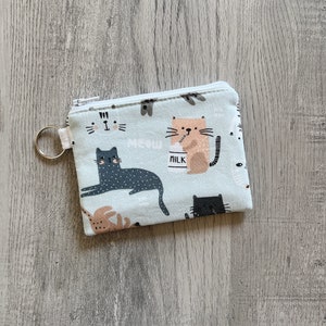 May include: A light blue zippered pouch with a white zipper pull and a silver key ring. The pouch is decorated with a repeating pattern of cats drinking milk, with the word "MEOW" printed on the pouch.