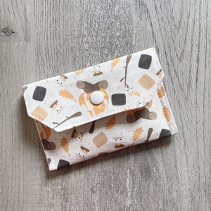 May include: A small, white fabric wallet with a flap closure, featuring a repeating pattern of marshmallows, campfires, and autumn leaves in shades of brown, orange, and tan. The wallet is secured with a white button.