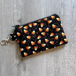 May include: Black zippered pouch with a candy corn pattern. The pouch features a repeating pattern of orange, yellow, and white candy corn on a black background. A silver-colored clasp is attached to the side. The pouch is likely used for carrying small items.
