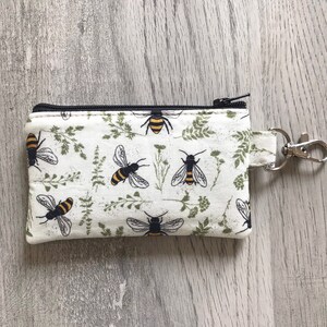 May include: A white zippered pouch with a black zipper and a black and yellow bee pattern. The pouch has a silver key ring attached to it.