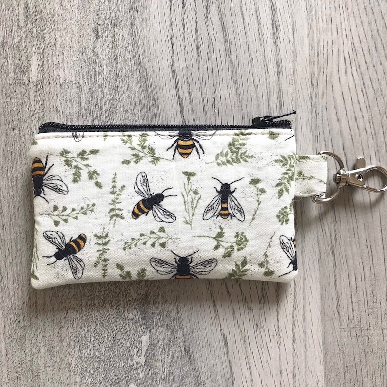 May include: A white zippered pouch with a black zipper and a black and yellow bee pattern. The pouch has a silver key ring attached to it.