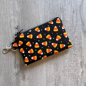 May include: A black zippered pouch with a candy corn pattern. The pouch features a repeating pattern of orange, yellow, and white candy corn on a black background. A silver-colored clasp is attached to the side.
