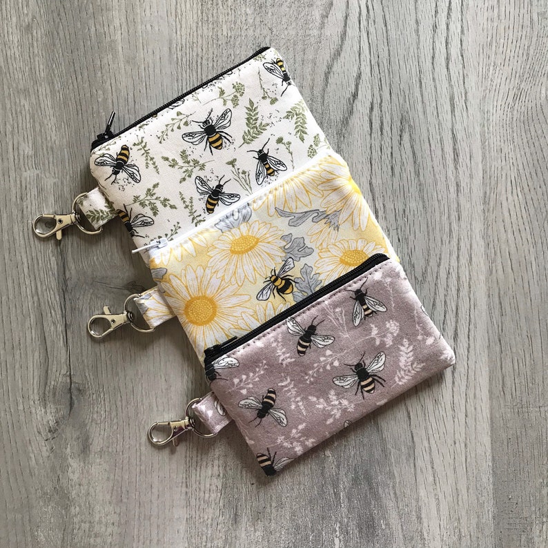 May include: Three zippered pouches with floral and bee designs. The pouches are stacked on top of each other and have black zipper pulls. The top pouch is white with green leaves and bees, the middle pouch is yellow with white flowers and bees, and the bottom pouch is purple with white flowers and bees.