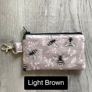 May include: A small zippered pouch with a key ring, featuring a light brown fabric with a repeating pattern of black bees and floral designs. The text "Light Brown" is printed below the pouch.