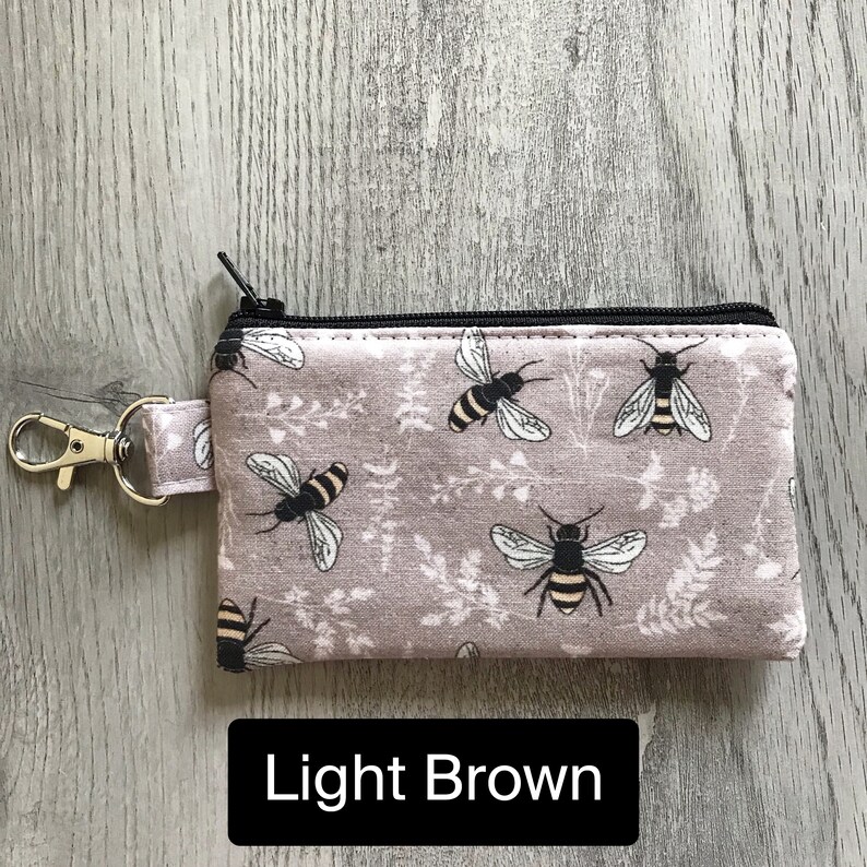 May include: A small zippered pouch with a key ring, featuring a light brown fabric with a repeating pattern of black bees and floral designs. The text "Light Brown" is printed below the pouch.