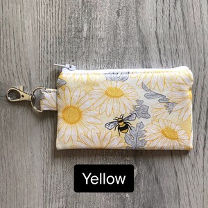 May include: A small yellow zippered pouch with a white daisy and bee print. The pouch has a silver key ring attached.