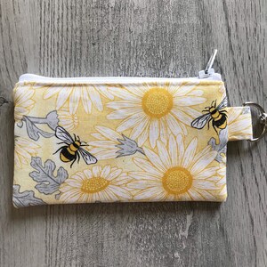 May include: A yellow and white zippered pouch with a daisy print and two black and yellow bees. The pouch has a white zipper and a white loop with a metal ring.
