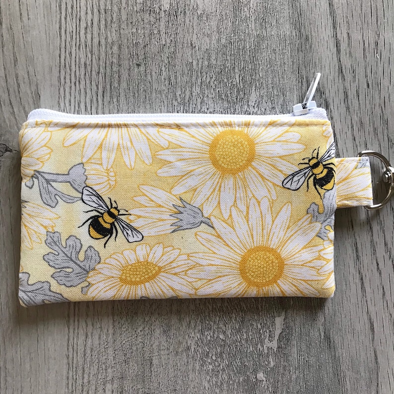 May include: A yellow and white zippered pouch with a daisy print and two black and yellow bees. The pouch has a white zipper and a white loop with a metal ring.