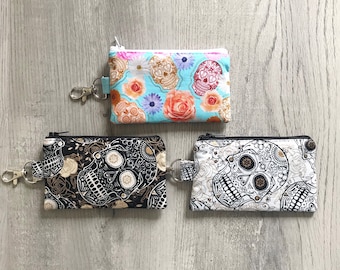 May include: Three small zippered coin purses with key rings. One purse has a light blue background with floral and skull patterns. The second purse has a black background with skull and floral patterns. The third purse has a white background with skull patterns.