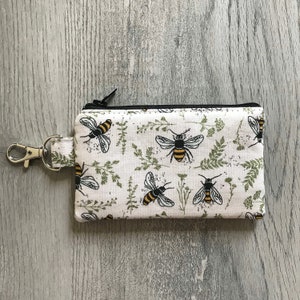 Peut inclure: Une pochette zippée blanche avec une fermeture éclair noire et un porte-clés noir. La pochette présente un motif répétitif d'abeilles et de feuillage vert sur fond blanc.