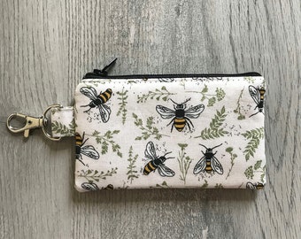 May include: A white zippered pouch with a black zipper and a black key chain. The pouch has a repeating pattern of bees and green foliage on a white background.