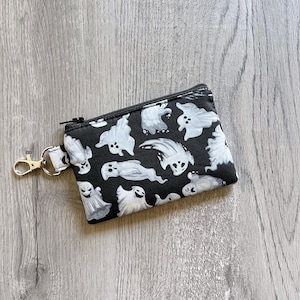 May include: Black zippered coin purse with a white ghost pattern. The small pouch has a gold-colored clasp and a black zipper. The ghosts are in various poses and sizes, creating a spooky Halloween theme.
