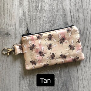 May include: Tan zippered pouch with a bee print. The pouch has a black zipper and a silver key ring.